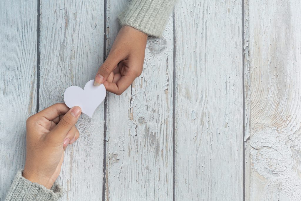 deux mains sur une table en bois blanche se donnant un coeur en papier